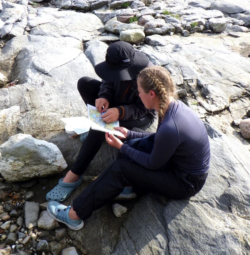 Two people are sitting on a rocky surface, possibly a large boulder or a rocky outcrop. One person is wearing a dark hat and appears to be pointing at a map, while the other person is looking at the map. Both are dressed in casual clothing suitable for outdoor activities. The surrounding environment consists of more rocks and stones, suggesting a natural, possibly coastal or mountainous setting.